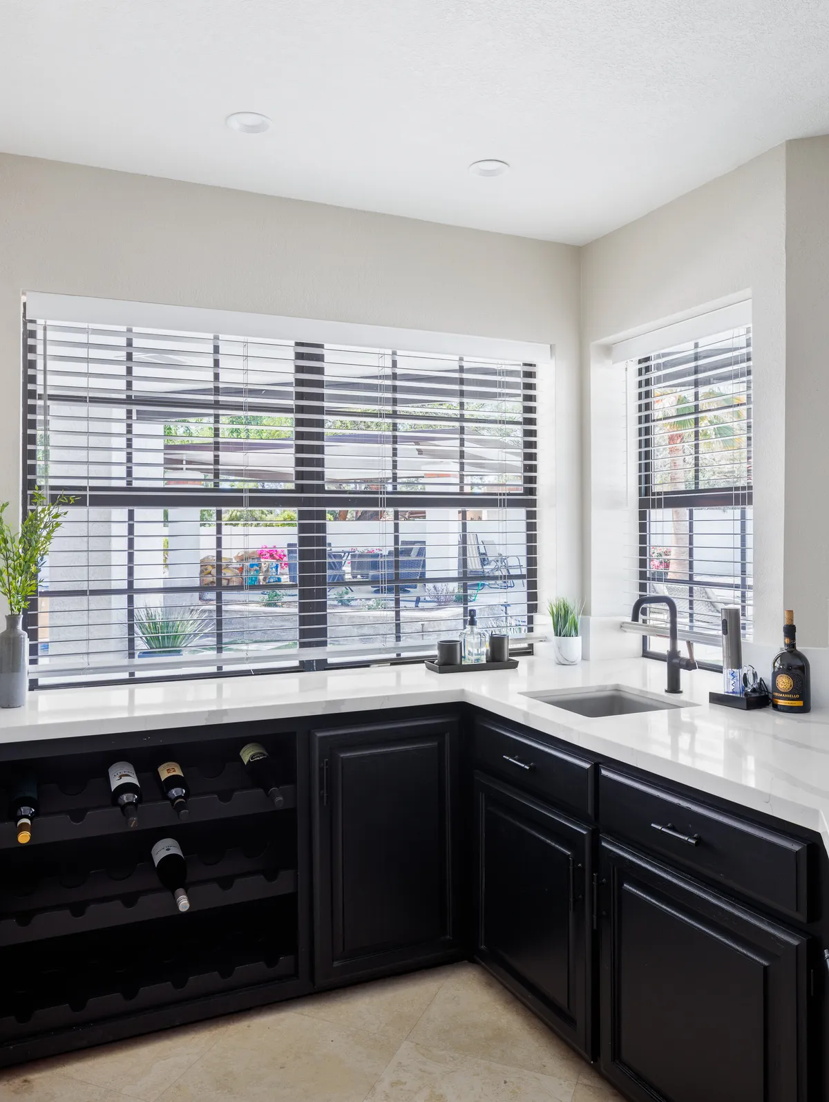 A sophisticated wet bar area featuring a modern wine rack filled with bottles, a sink with a sleek faucet, and a bright white countertop. Natural light pours in through the window blinds, suggesting an inviting spot for mixing drinks or wine tasting with guests.