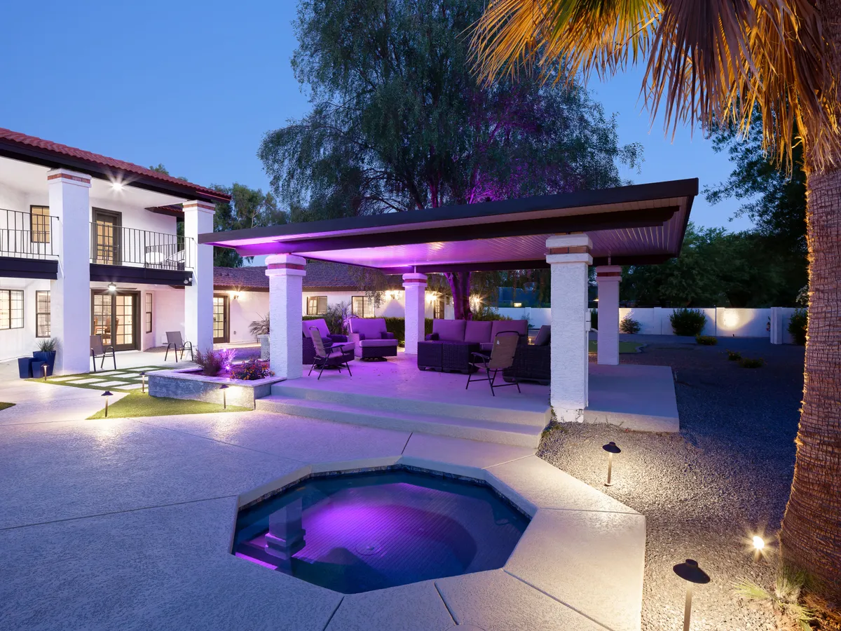 A stylish backyard scene illuminated for evening enjoyment, featuring a pool with purple lighting, a covered seating area under a gazebo, and a contemporary two-story building framing the space. The ambiance is both tranquil and festive, ideal for gatherings or quiet relaxation.