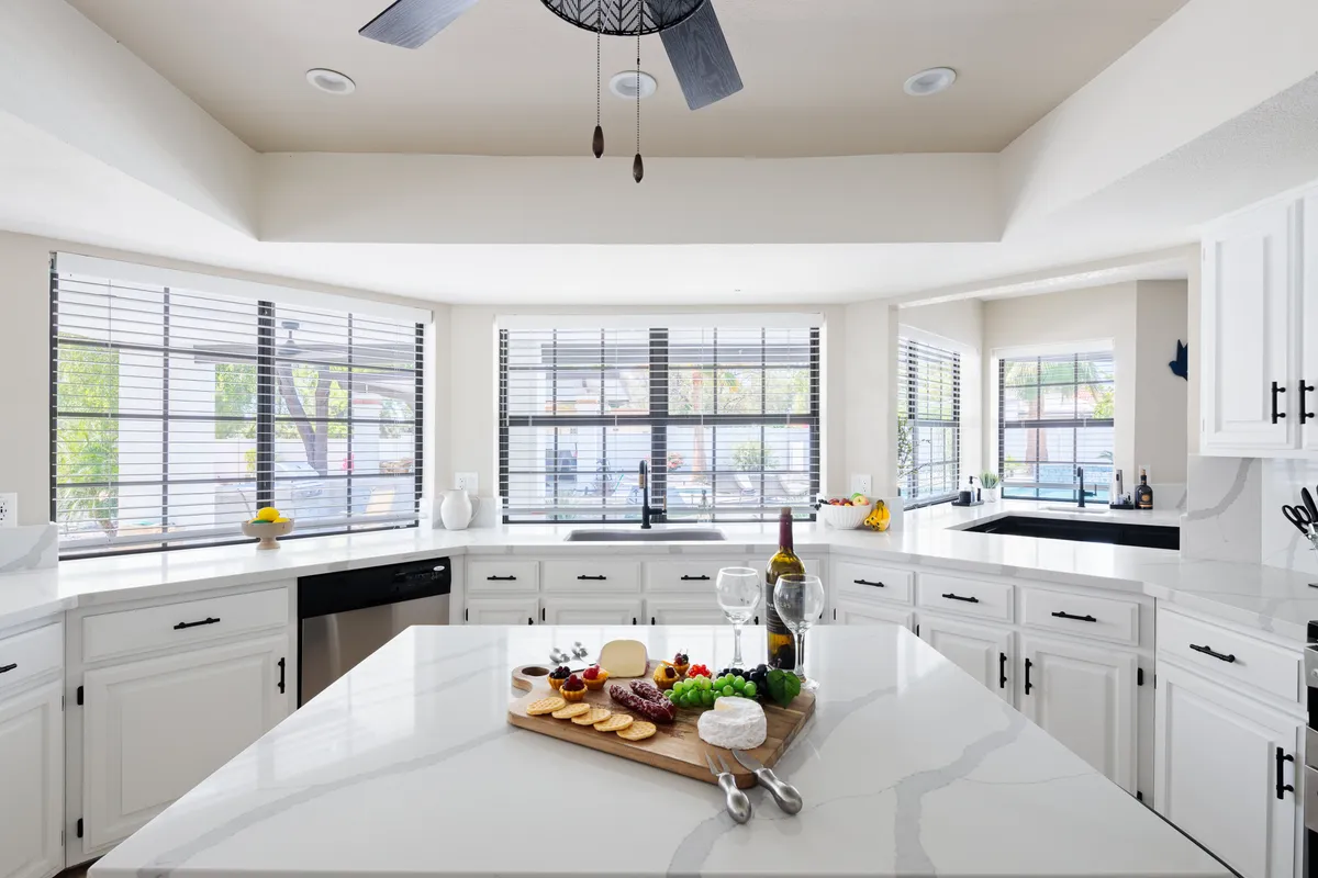The scene features a central kitchen island adorned with a charcuterie board, wine bottles, and glasses, under warm natural light from large windows. The atmosphere is lively and inviting, emphasizing shared meals and celebratory occasions in the heart of the home.